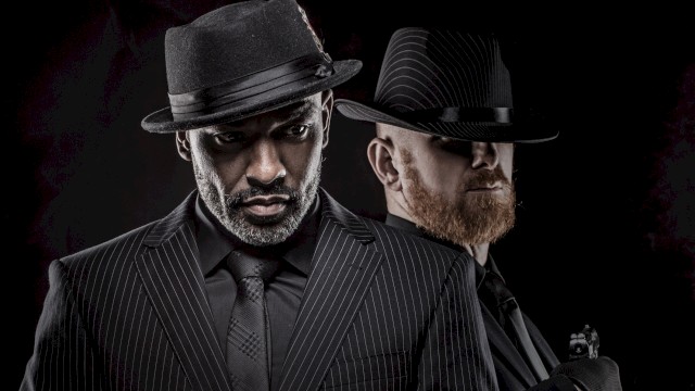 Two men dressed in black suits and hats,pose dramatically against a dark background in a cinematic style.