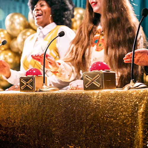 People are having fun, pressing red buttons at a lively event decorated with golden balloons and a shimmering tablecloth.