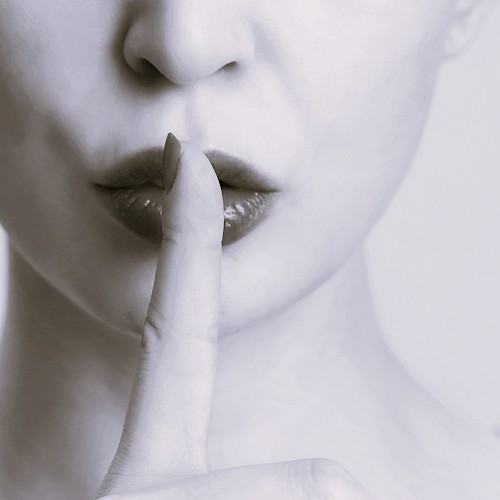 A close-up of a person with their finger on their lips, signaling to keep a secret or stay silent, in monochrome tones.