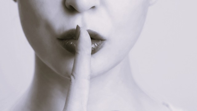 A close-up of a person with their finger on their lips, signaling to keep a secret or stay silent, in monochrome tones.