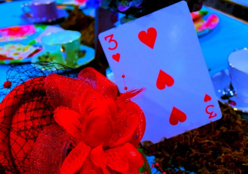 The image features a three of hearts playing card, decorated with red fabric and feathers, among colorful tableware and decorations.