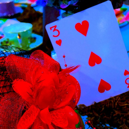 The image features a three of hearts playing card, decorated with red fabric and feathers, among colorful tableware and decorations.
