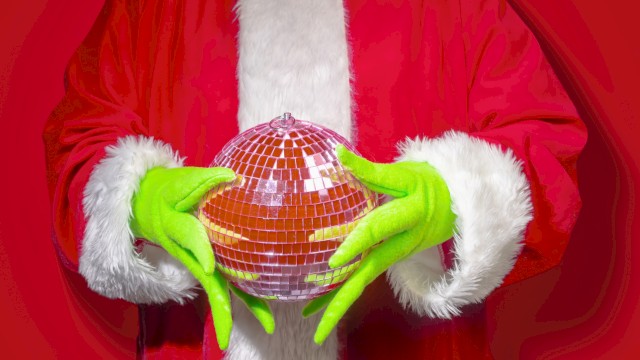 A person dressed as Santa, wearing green gloves, holding a disco ball with a red background.