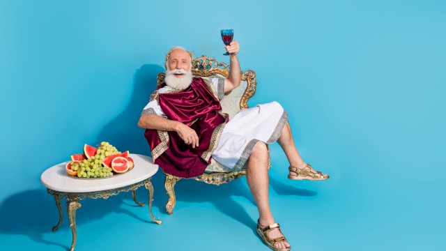 Older man dressed as a Roman emperor, sitting on a vintage chair, holding a glass of red wine with a tray of fruit beside him.