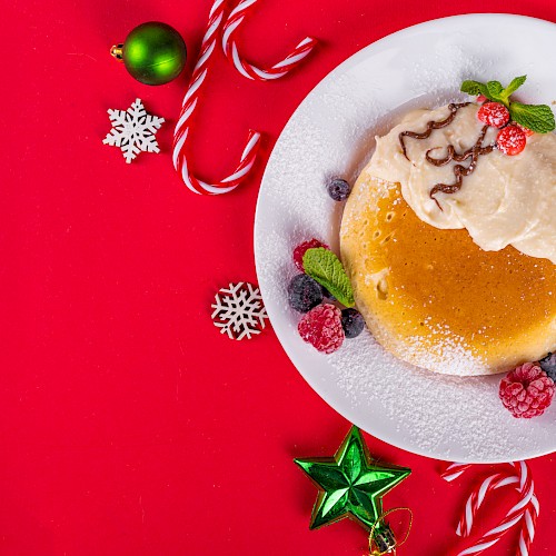 A decorated Xmas pudding on a white plate, surrounded by holiday ornaments, candies, and snowflakes on a red background.