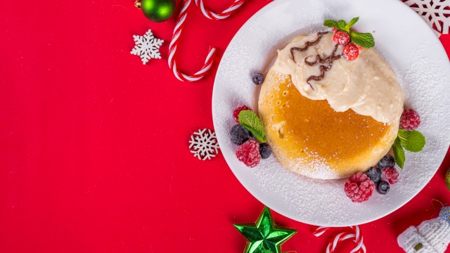 A decorated Xmas pudding on a white plate, surrounded by holiday ornaments, candies, and snowflakes on a red background.