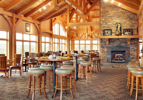 A spacious wooden lodge interior features a stone fireplace, high ceilings, numerous tables with stools, and large windows allowing natural light in.