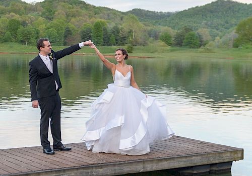 A bride and groom stand on a wooden dock by a lake, with the groom twirling the bride. The background features greenery and rolling hills.
