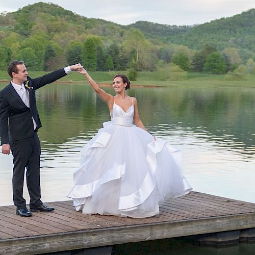 A bride and groom stand on a wooden dock by a lake, with the groom twirling the bride. The background features greenery and rolling hills.