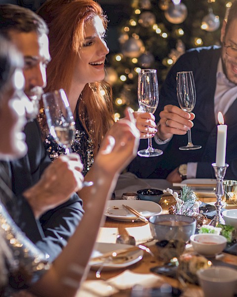 A group of people celebrating at a dinner table, holding champagne glasses, with a Christmas tree in the background.