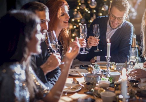 A group of people celebrating at a dinner table, holding champagne glasses, with a Christmas tree in the background.