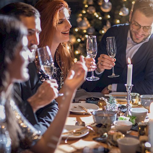 A group of people celebrating at a dinner table, holding champagne glasses, with a Christmas tree in the background.