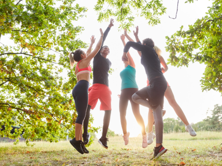 A group of five people wearing athletic clothing is jumping and high-fiving outdoors in a park, surrounded by green trees and sunlight at the end.