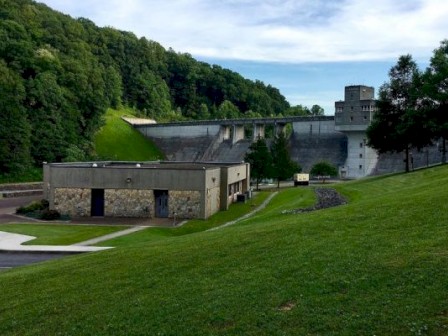 The image shows a dam with adjacent buildings surrounded by green landscape and trees, under a partly cloudy sky.