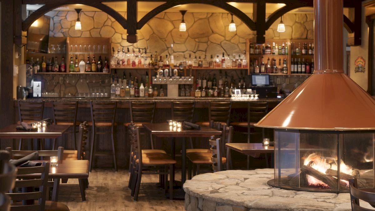 A cozy restaurant bar with stone and wood decor, featuring a central fireplace, wooden tables and chairs, and a well-stocked bar with various bottles.