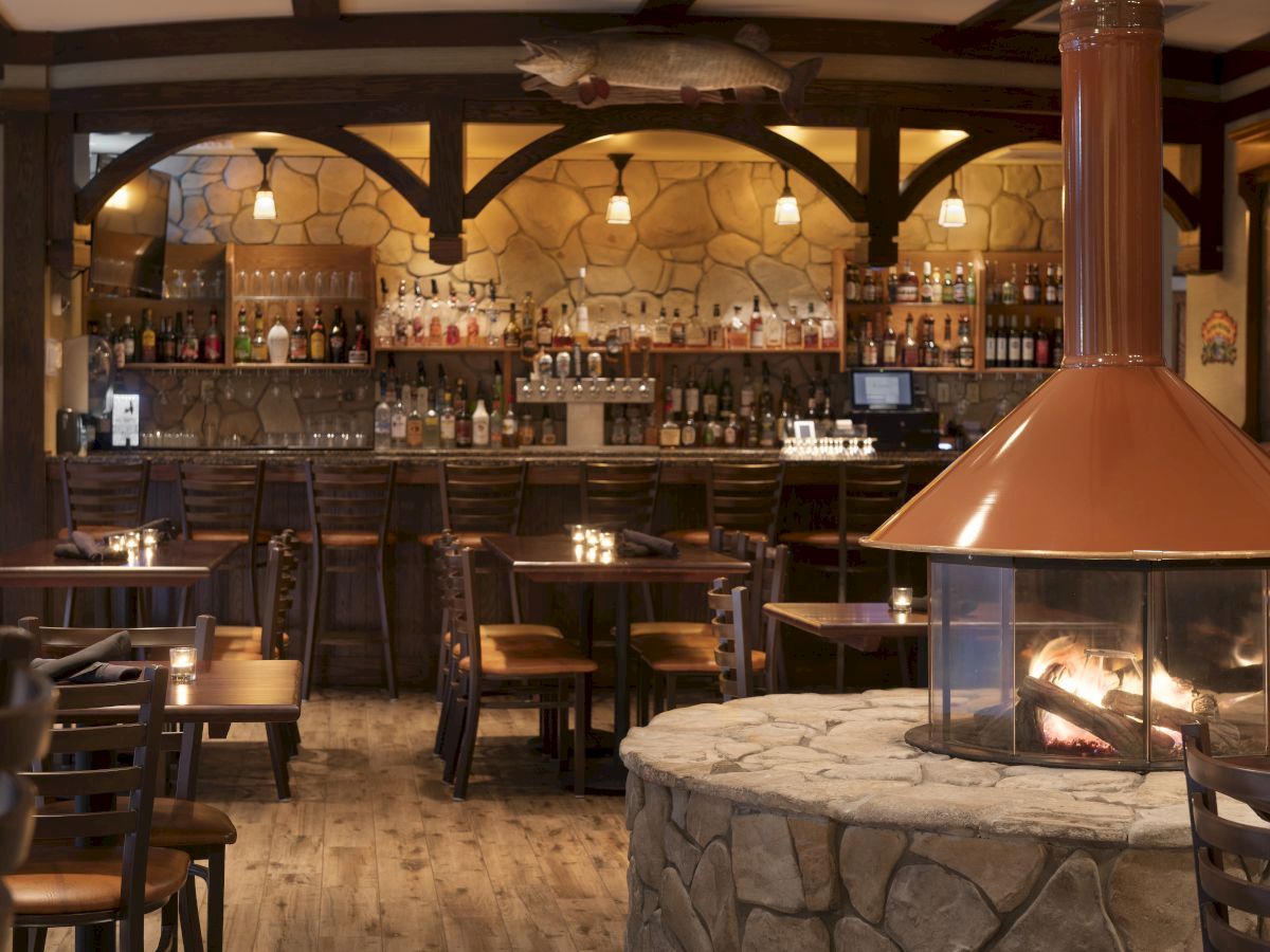 A cozy restaurant bar with stone and wood decor, featuring a central fireplace, wooden tables and chairs, and a well-stocked bar with various bottles.