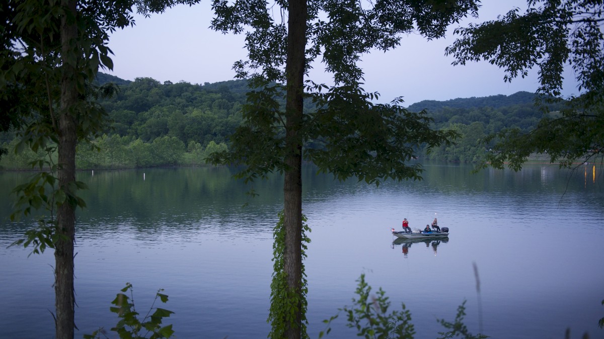 A serene lake surrounded by trees, with two people on a small boat in the center of the water, set against a background of hills.