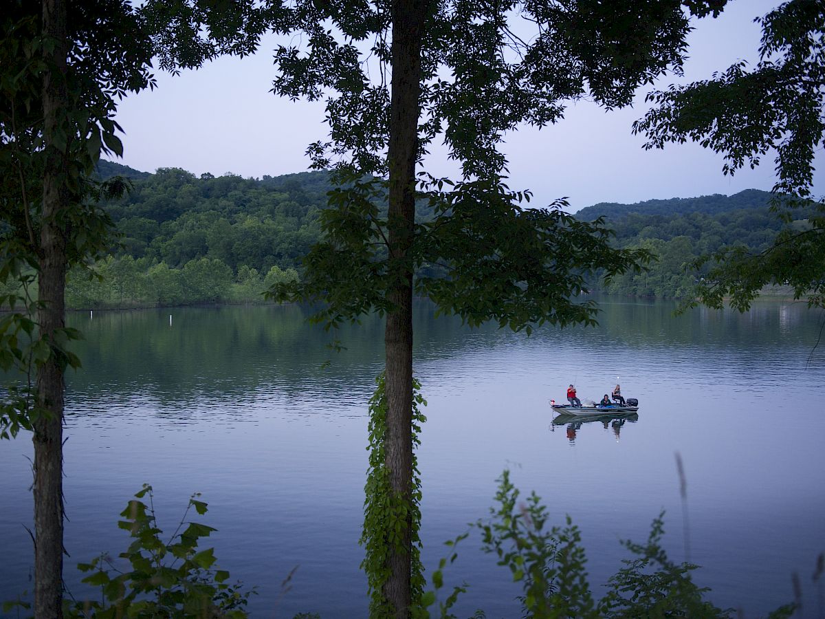 A serene lake surrounded by trees, with two people on a small boat in the center of the water, set against a background of hills.