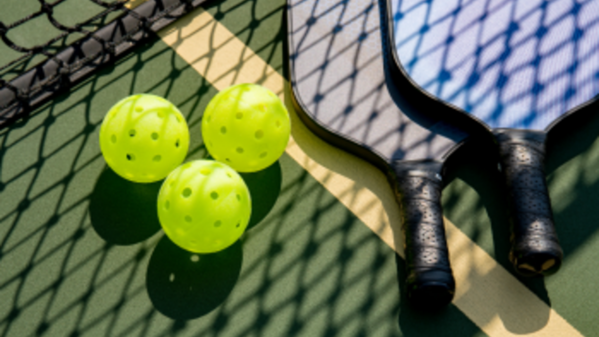 The image shows two pickleball paddles, three yellow perforated pickleball balls, and part of a net on an outdoor court.