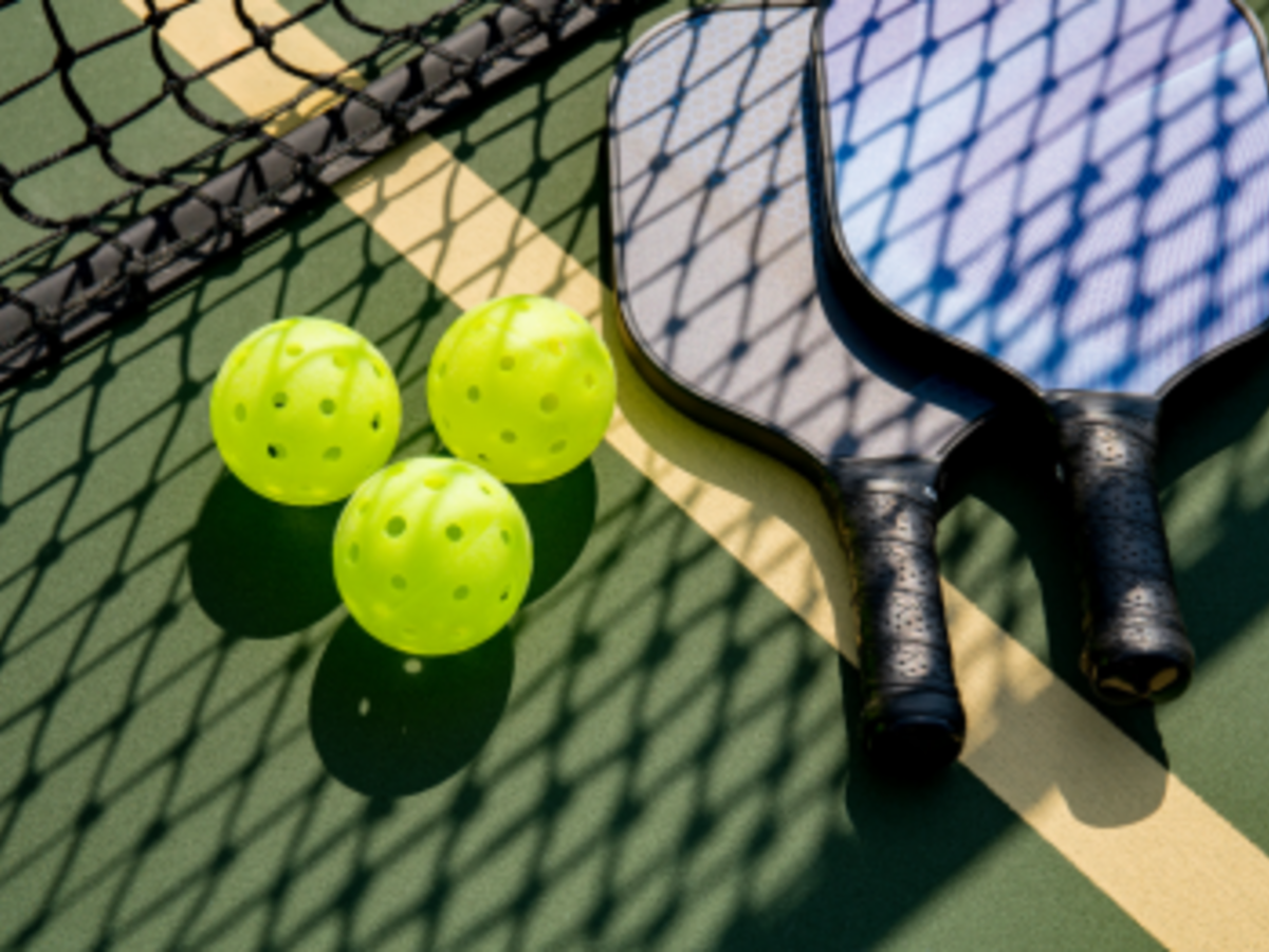 The image shows two pickleball paddles, three yellow perforated pickleball balls, and part of a net on an outdoor court.