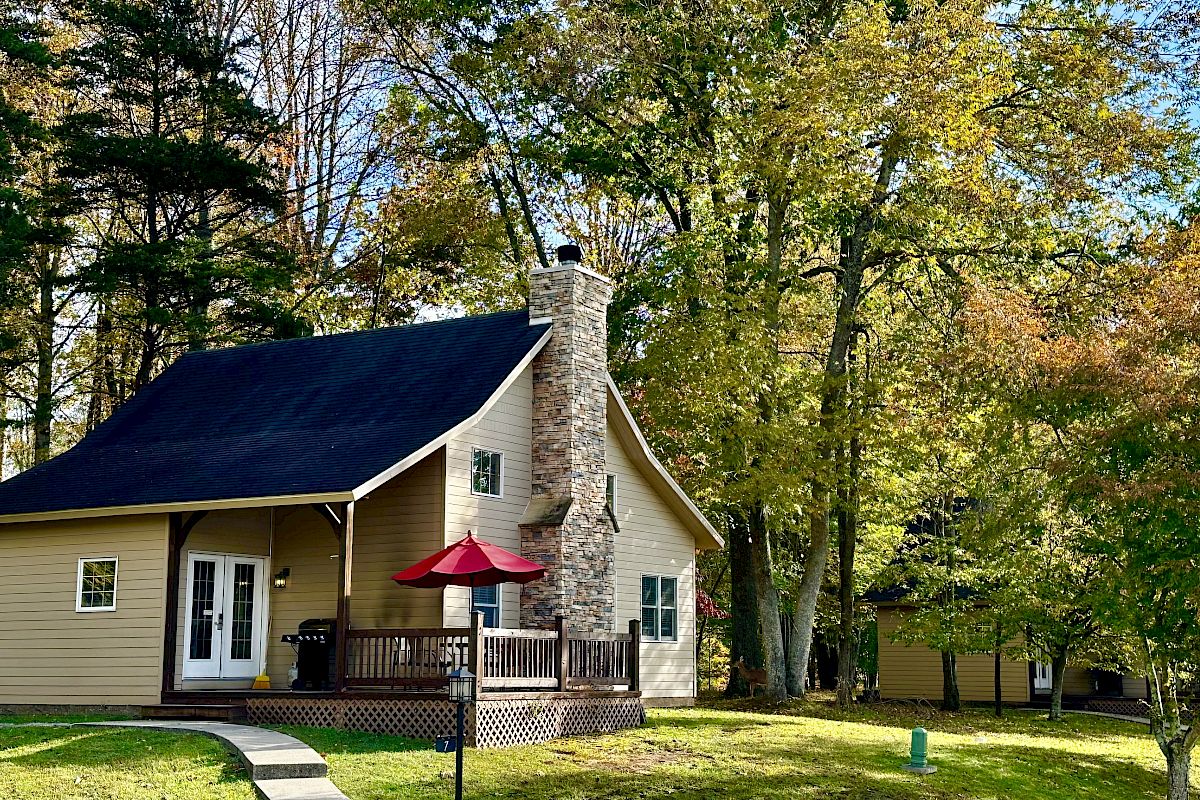 A cozy cottage with a stone chimney, surrounded by trees and grass, featuring a red umbrella-covered patio in front.