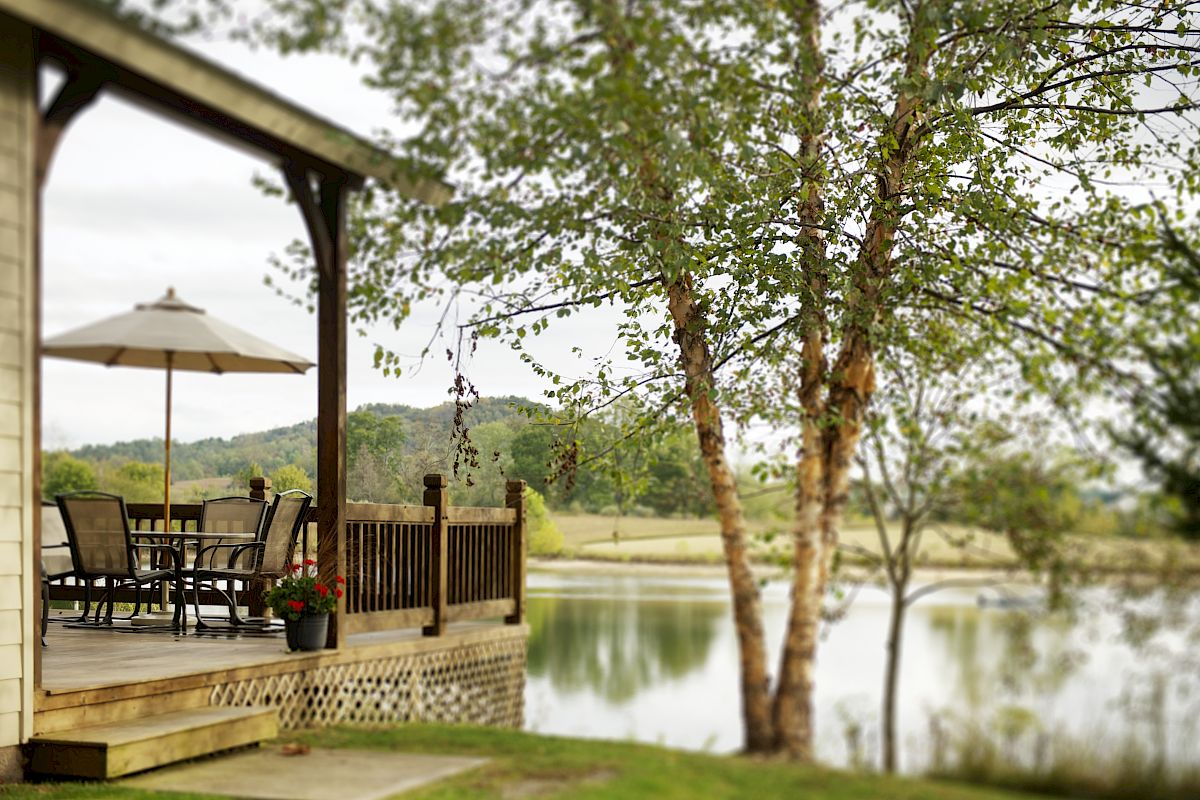 A cozy porch with a table and chairs under an umbrella, near a calm lake and surrounded by trees, offering a peaceful outdoor setting.