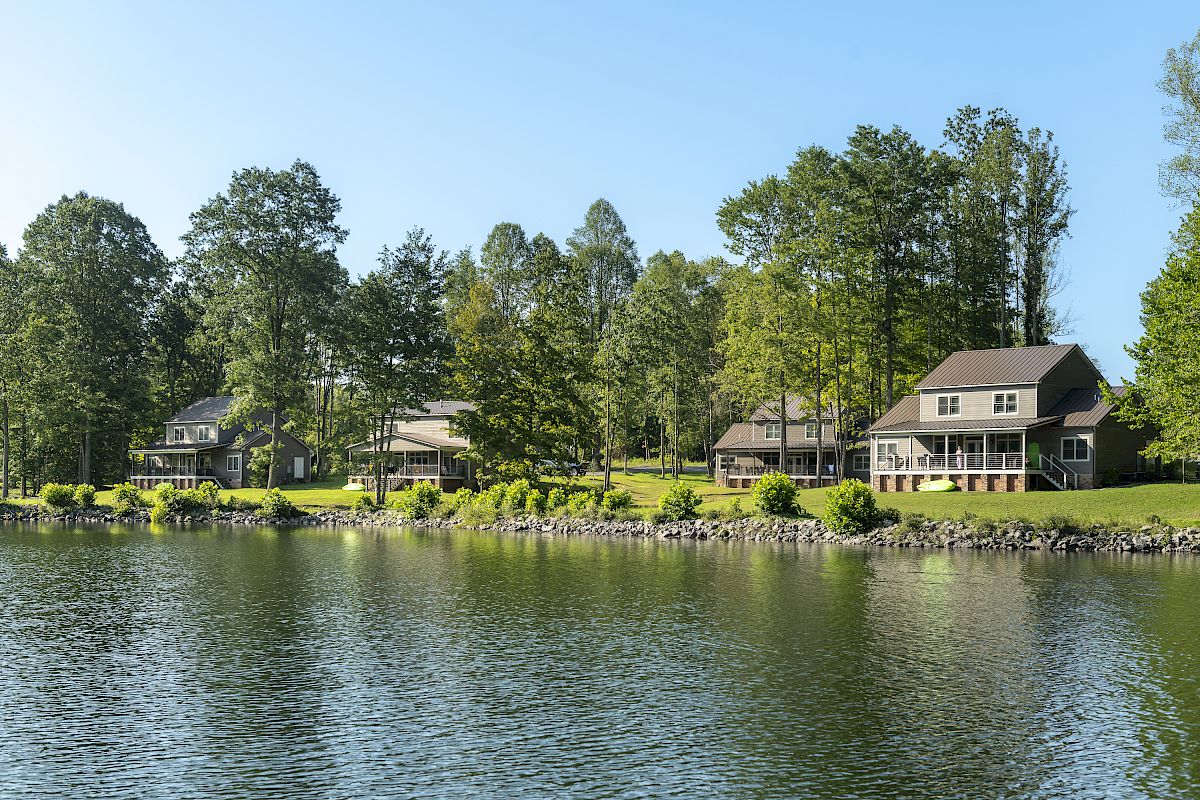 The image shows lakeside houses with green lawns and trees, reflecting off the water under a clear blue sky.