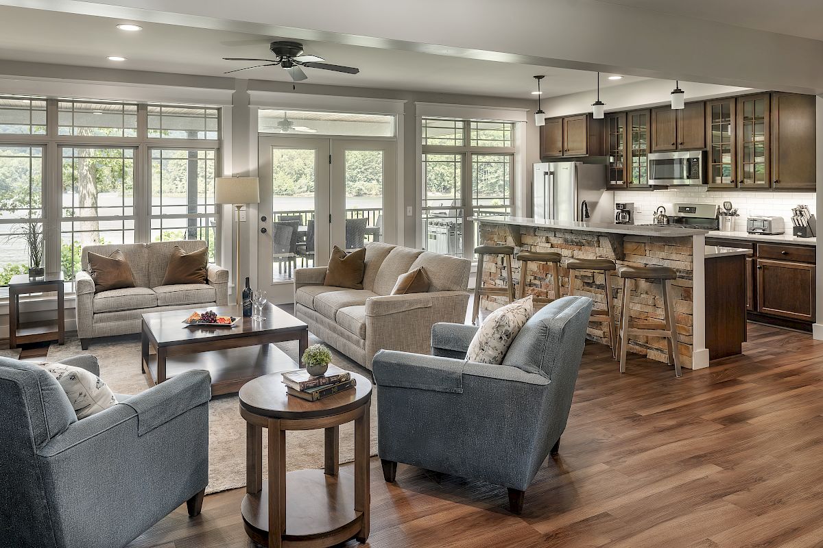 A spacious, modern living room and kitchen area with sofas, armchairs, a coffee table, kitchen island, and large windows overlooking a view.