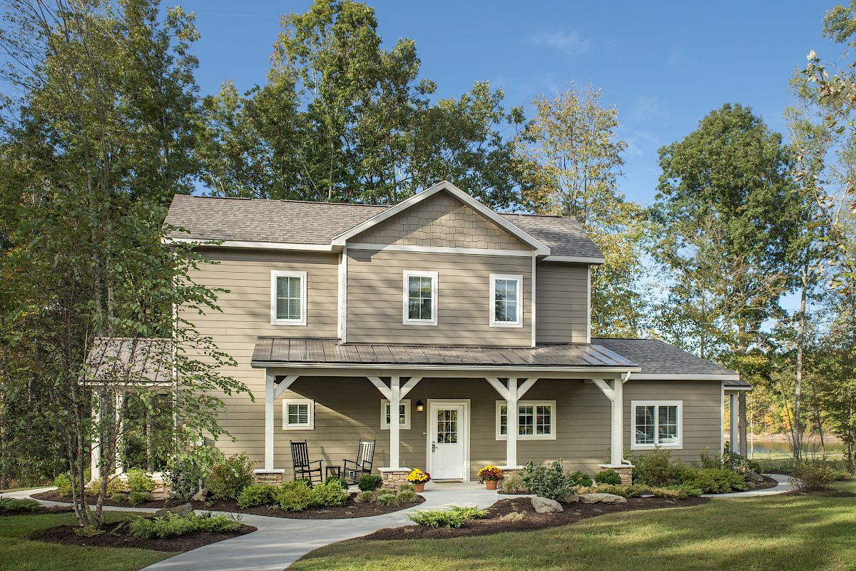 A two-story house with a covered porch is surrounded by trees, featuring a garden and a curved walkway leading to the front door.