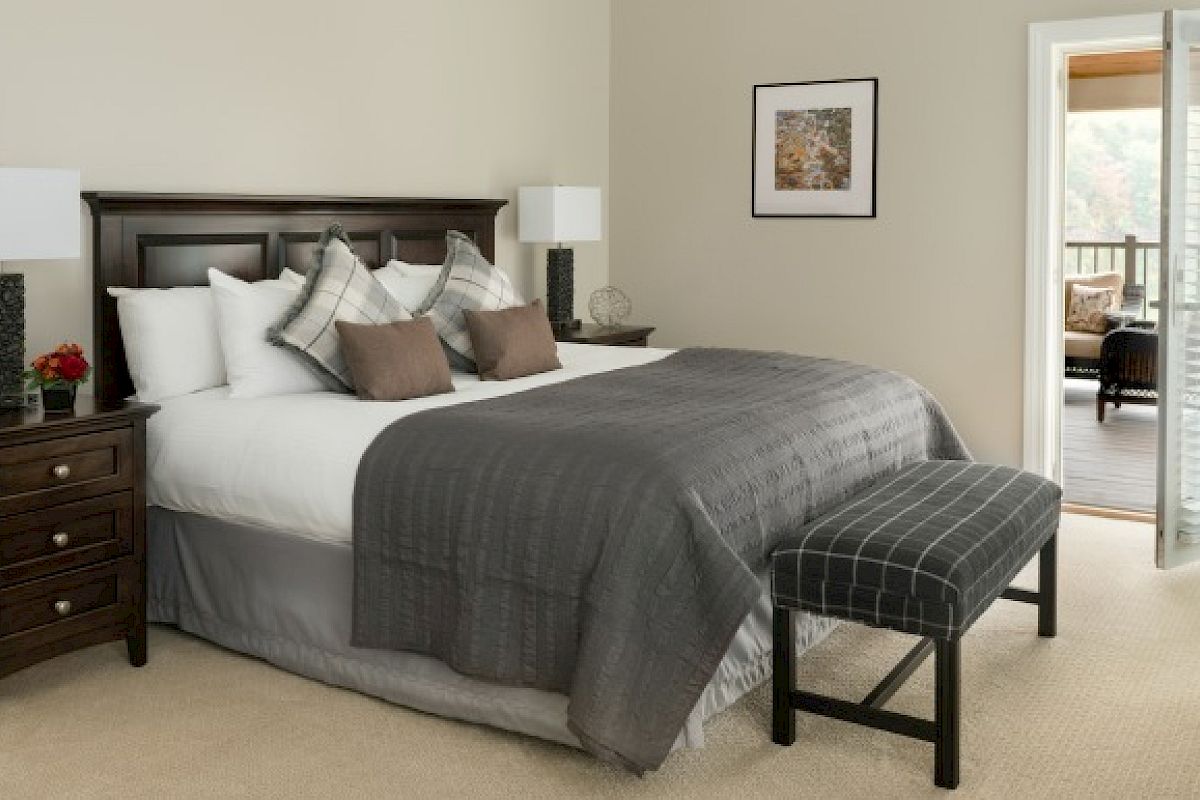A neatly arranged bedroom with a large bed, nightstands, lamps, a bench, wall art, and an open door leading to a balcony.