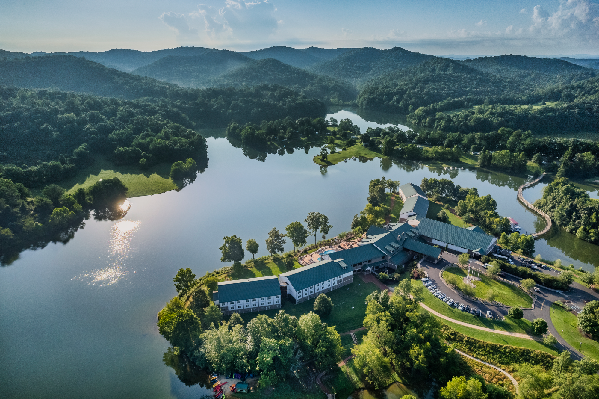 Aerial view of a lakeside resort surrounded by lush green forests and hills, with a winding road and parking area, all bathed in sunlight.