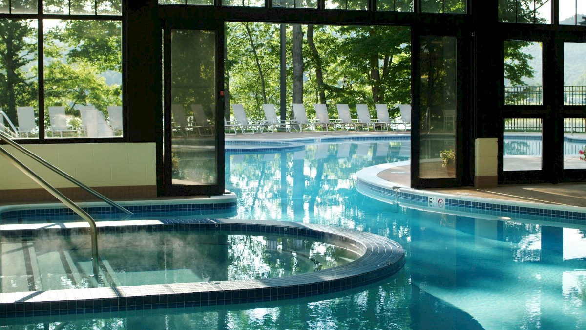 An indoor pool area with large glass doors opens to an outdoor pool surrounded by lush trees and lounge chairs, creating a serene environment.