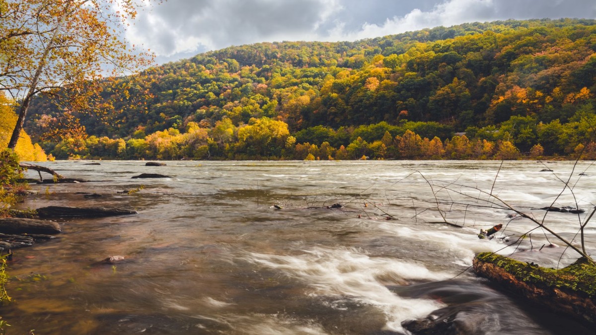 A river flows in the foreground, surrounded by autumn foliage. In the background, there are forested hills under a partly cloudy sky.