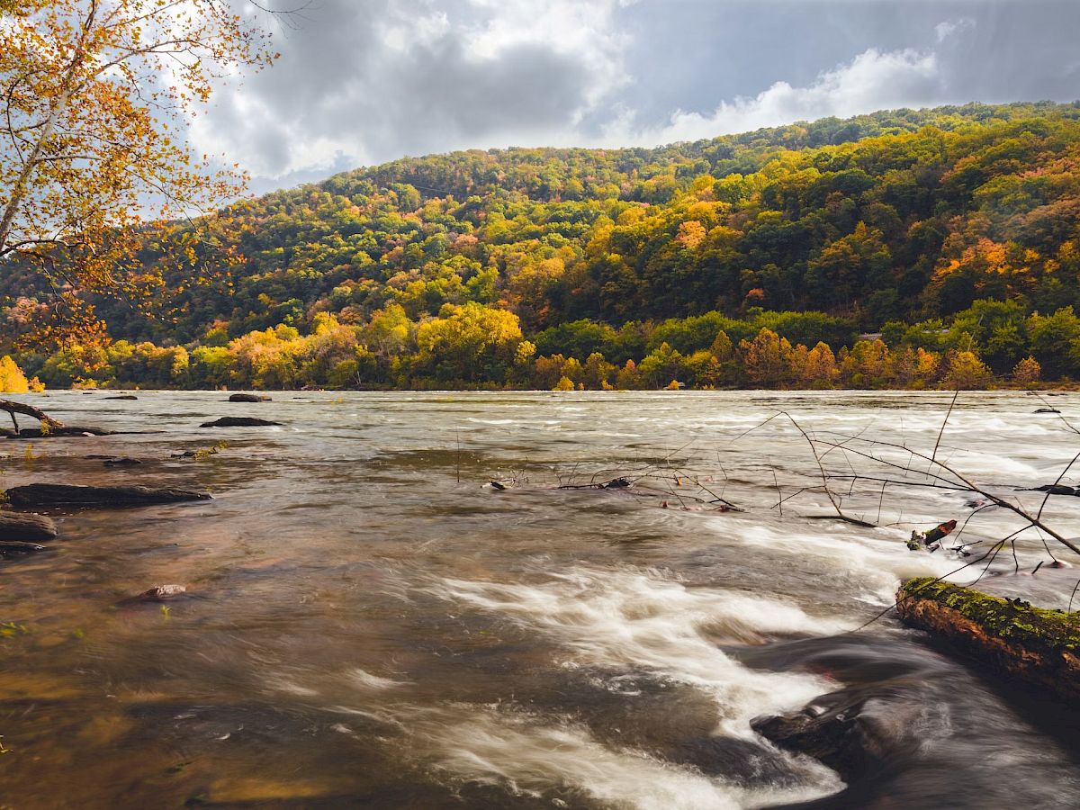 A river flows in the foreground, surrounded by autumn foliage. In the background, there are forested hills under a partly cloudy sky.