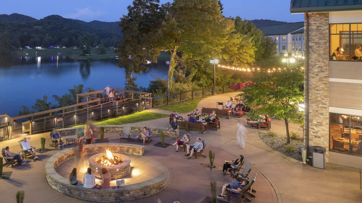 People gathered around a fire pit on a patio near a lake, surrounded by trees and outdoor seating at dusk.