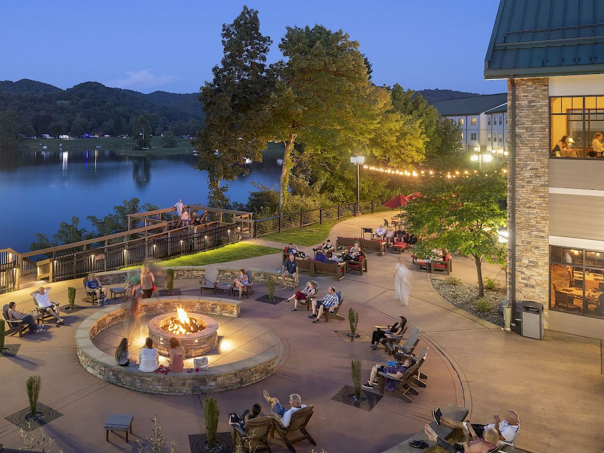 People gathered around a fire pit on a patio near a lake, surrounded by trees and outdoor seating at dusk.