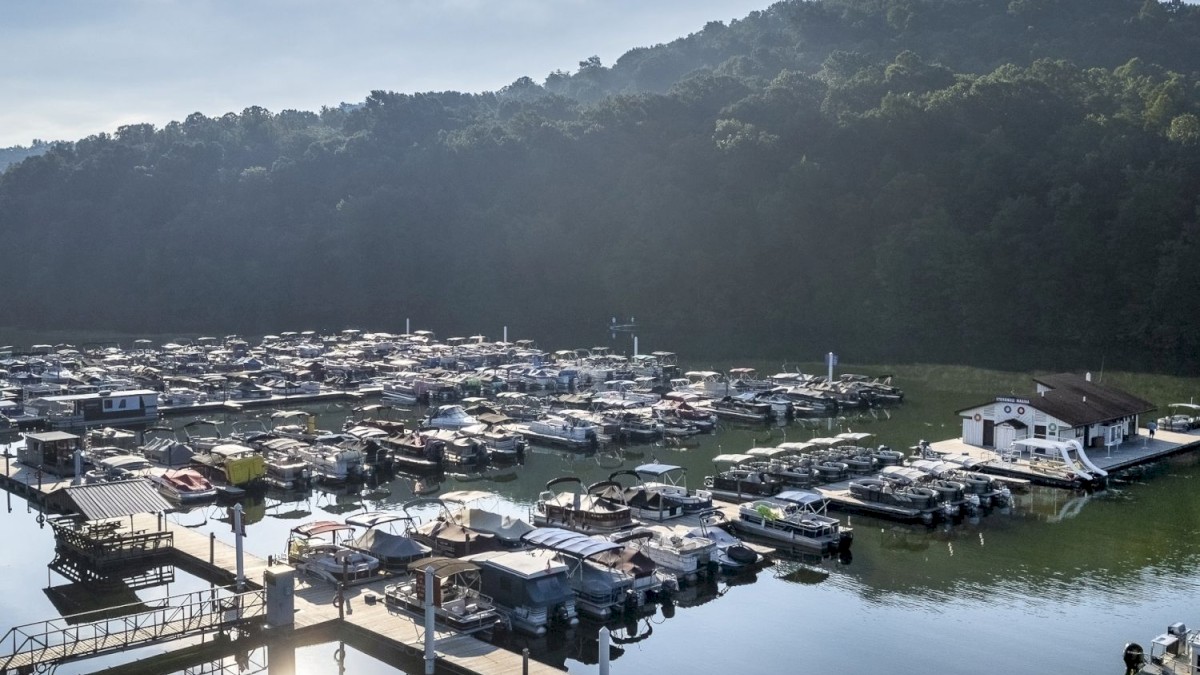 The image shows a harbor with numerous boats docked, calm water reflecting the sky, a hilly backdrop, and a bright sun overhead.