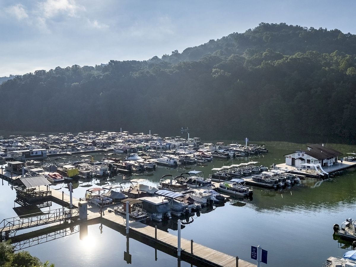 The image shows a harbor with numerous boats docked, calm water reflecting the sky, a hilly backdrop, and a bright sun overhead.