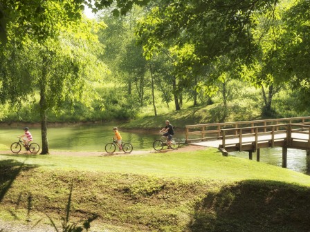 Three people are bicycling on a path near a wooden bridge beside a scenic lake surrounded by lush green trees and grass, ending the sentence.