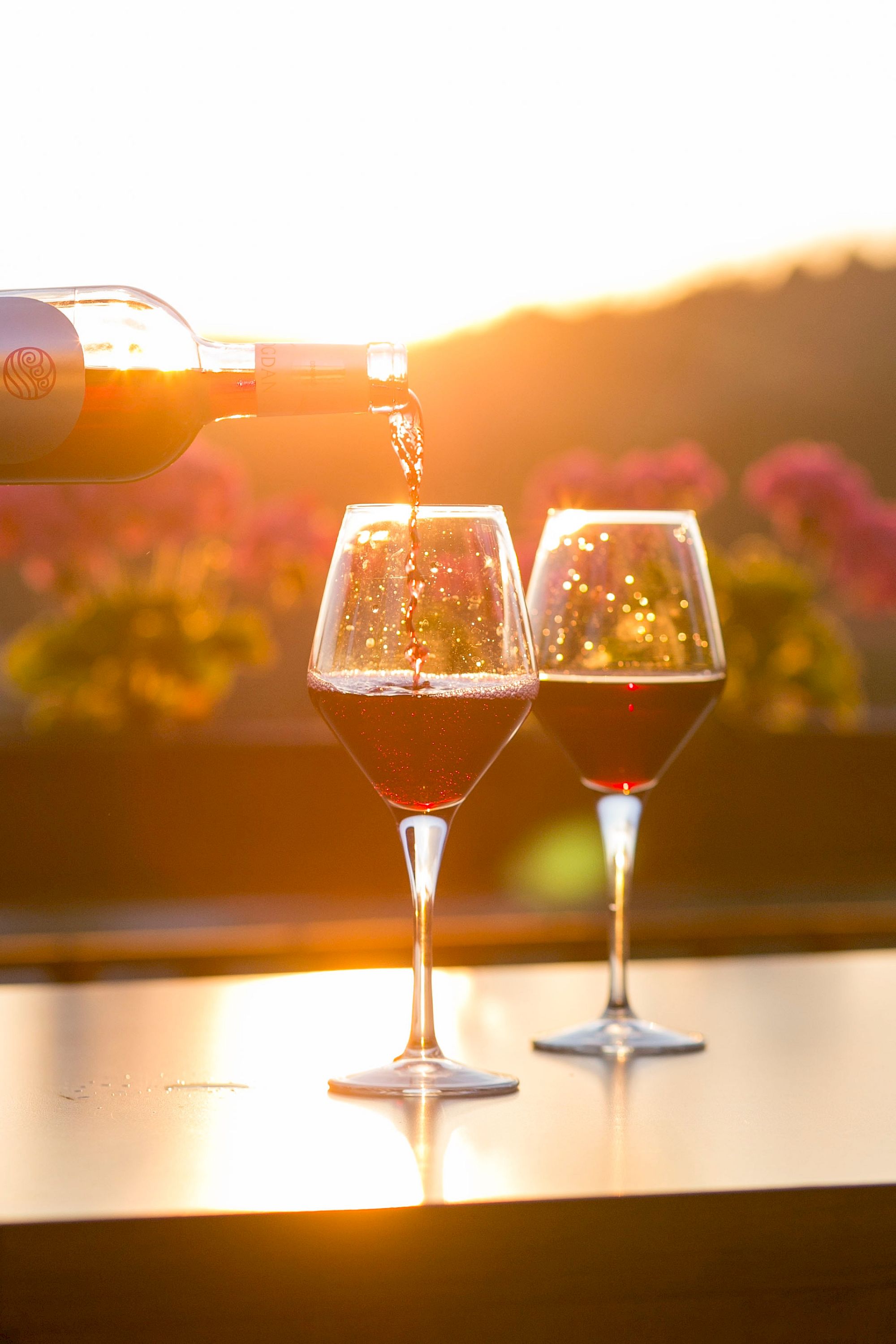 A bottle of wine pours into two glasses on a table at sunset, with a blurred background of flowers and hills.