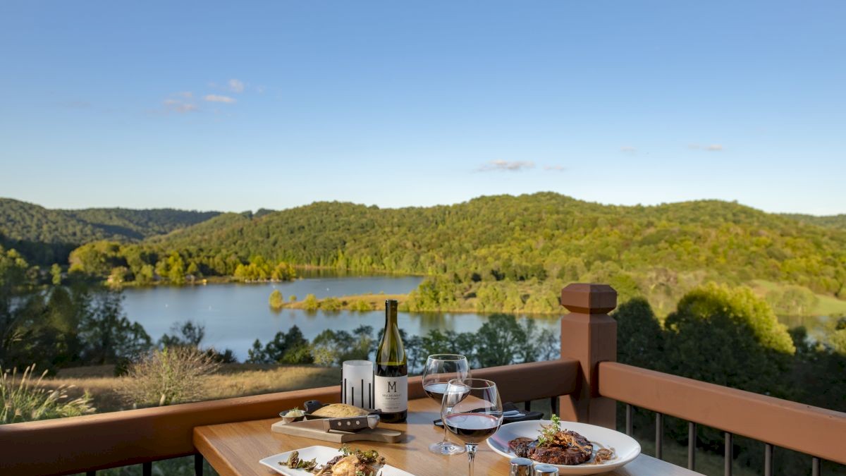 A wooden table on a balcony with dishes, wine, and glasses, overlooking a serene lake and lush green hills under a clear blue sky.