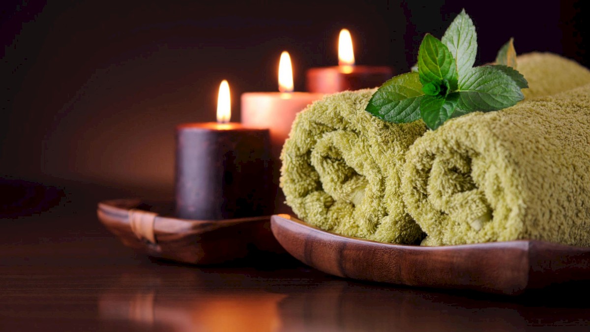 The image shows rolled green towels, lit candles, and a sprig of green leaves arranged on a wooden surface, creating a relaxing atmosphere in one sentence.