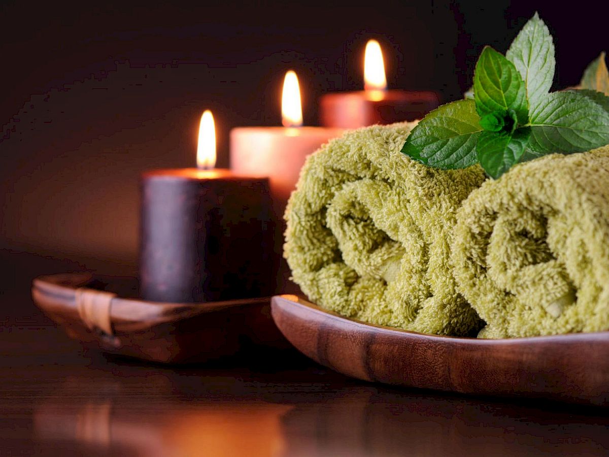 The image shows rolled green towels, lit candles, and a sprig of green leaves arranged on a wooden surface, creating a relaxing atmosphere in one sentence.