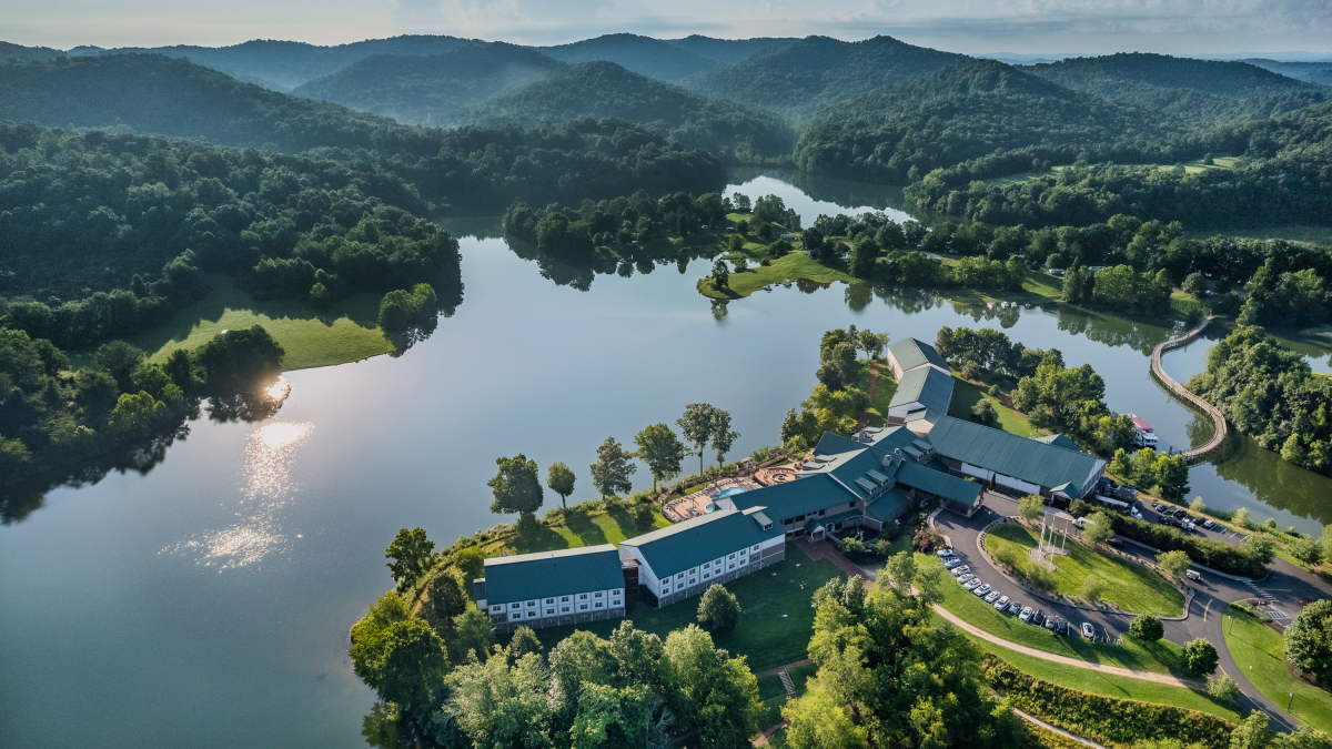 An aerial view of a large lodge or resort nestled near a lake with surrounding wooded hills and lush green landscape on a clear day.