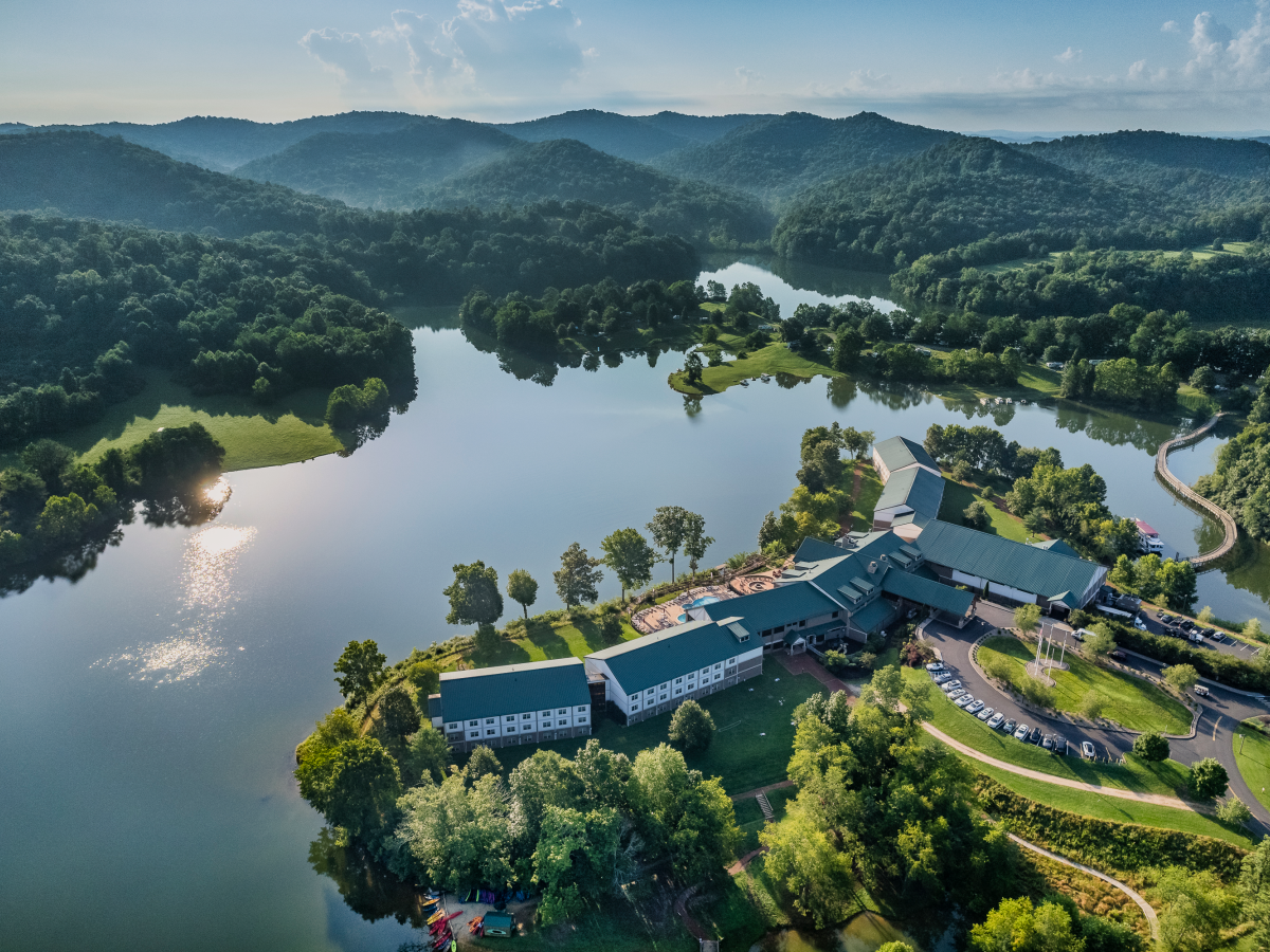 An aerial view of a large lodge or resort nestled near a lake with surrounding wooded hills and lush green landscape on a clear day.