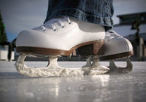 The image shows a pair of white ice skates being worn, positioned on the ice, possibly at a skating rink.