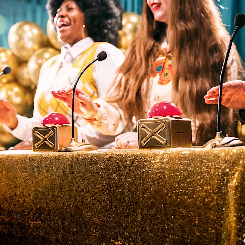 People are participating in a game show, with hands ready to press red buzzers, in front of a gold sequin tablecloth and golden balloons.