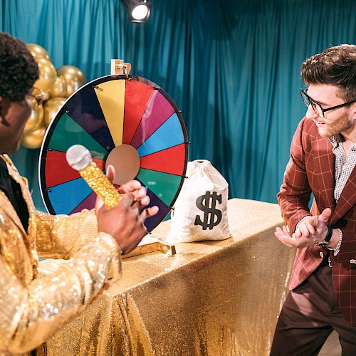 Two men are interacting on a game show set with a prize wheel and a money bag, with the host holding a microphone.