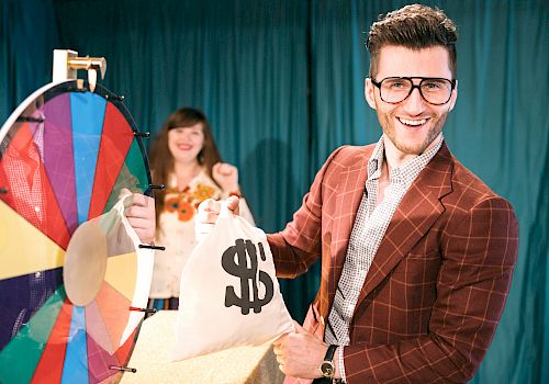 A man in a suit with glasses holds a money bag and smiles in front of a colorful spinning wheel, while a woman in the background looks pleased.