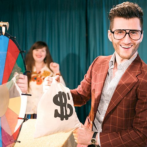 A man in a suit with glasses holds a money bag and smiles in front of a colorful spinning wheel, while a woman in the background looks pleased.